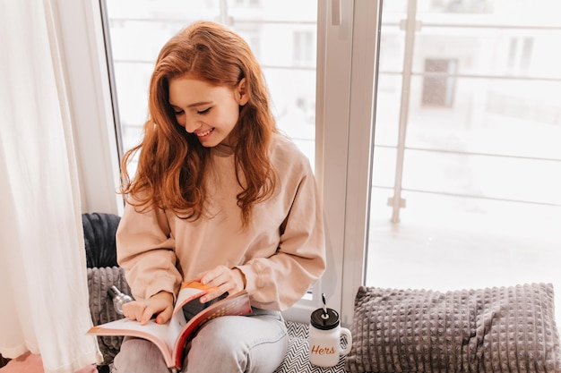 Enthousiaste roodharige vrouw die tijdschrift leest Binnenportret van verlegen gembermeisje met boek