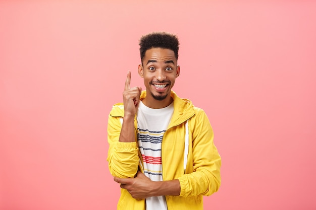 Enthousiaste tevreden afro-amerikaanse man die suggestie toevoegt om wijsvinger op te steken in eureka-gebaar en vreugdevol glimlacht terwijl hij interessante uitvinding of theorie bespreekt die breed over roze muur glimlacht