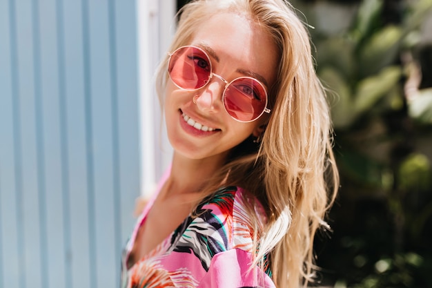 Enthousiaste vrouw in trendy roze bril op zoek naar camera met prachtige glimlach.