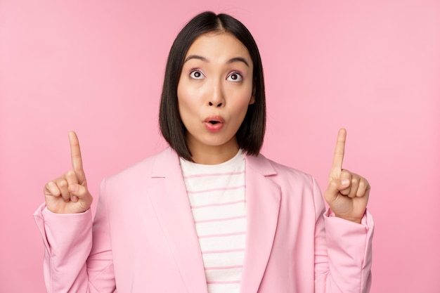 Enthousiaste zakelijke werknemer aziatische zakenvrouw die met de vingers omhoog wijst en glimlacht en een advertentielogo laat zien dat over een roze achtergrond staat