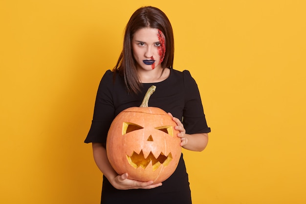 Gratis foto ernstig meisje met boze gelaatsuitdrukking die zich met pompoen in handen in studio bevindt die op geel wordt geïsoleerd, aantrekkelijk wijfje met bloedige wond op haar gezicht, halloween-concept.