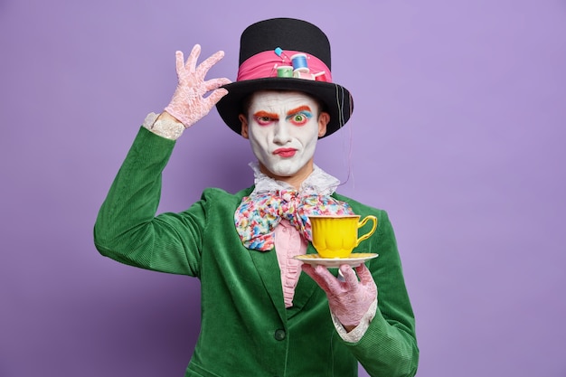 Ernstig mysterieus mannelijk personage uit wonderland fronst gezicht houdt hand op hoed drinkt thee op feestjurken voor halloween doet alsof hij een gekke hoedenmaker is heeft kleurrijke make-up geïsoleerd over paarse muur