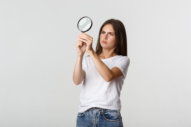 Ernstig ogende aantrekkelijke jonge vrouw op zoek naar iets, kijkend door vergrootglas, wit.