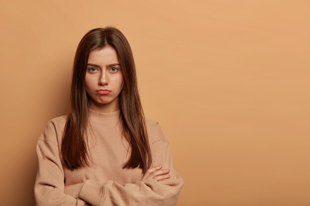 Ernstig uitziende beledigde Europese vrouw portretteert de lippen, houdt de handen gekruist, heeft een sombere, boos uitdrukking, klaagt, draagt een bruine sweater