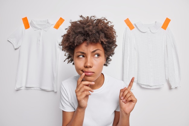 Ernstige aantrekkelijke vrouw met krullend haar wijst wijsvinger toont kleding gepleisterd aan witte muur gekleed in casual t-shirt poses binnen Vrouwelijk model adverteert kledingstukken Kijk hier eens naar