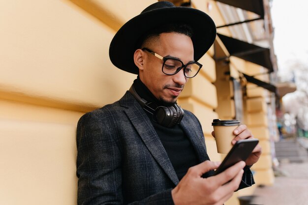 Ernstige Afrikaanse kerel die nieuws op internet leest terwijl hij koffie drinkt. Buiten foto van geconcentreerde zwarte jongeman in stijlvolle hoed staande met telefoon en latte in de buurt van gebouw.