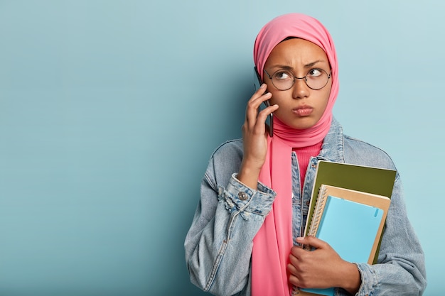 Ernstige Arabische vrouw belt via de mobiele telefoon, kijkt opzij, heeft een norse gezichtsuitdrukking en draagt een ronde bril