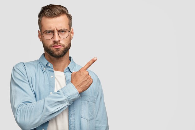 Ernstige bebaarde man met een strikte uitdrukking, trekt ergens je aandacht naar, gekleed in een blauw shirt, wijst richting ergens