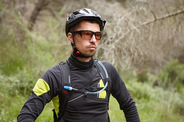 Ernstige blanke mannelijke wielrenner dragen van beschermende helm en bril moe voelen na intensieve fietstraining buitenshuis in bos op zijn booster fiets