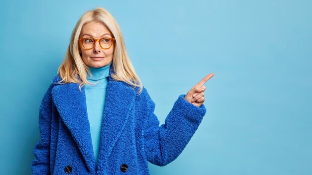 Ernstige blonde veertig jaar oude blanke vrouw heeft nieuwsgierige uitdrukking en geeft weg op lege ruimte draagt optische bril stijlvolle bontjas.