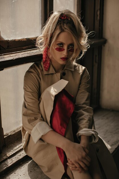 Ernstige blonde vrouw met paardenstaart en heldere lippen in rode zonnebril en beige trenchcoat zit op de vensterbank en kijkt in de camera bij de ramen