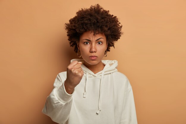 Ernstige boze etnische vrouw bedreigt je met gebalde vuist, zegt dat ik je zal laten zien hoe je ongehoorzaam kunt zijn, draagt een casual witte hoodie, belooft te straffen