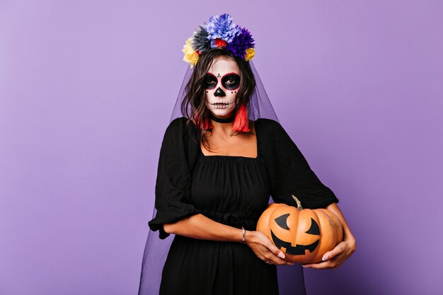 Ernstige bruinharige vrouw in sluier met halloween-pompoen. Binnen schot van prachtig meisje in dode bruid outfit met enge make-up.