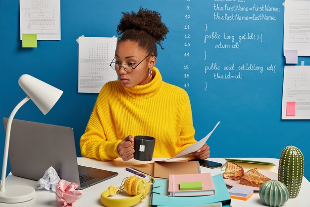 Ernstige donkerhuidige vrouw controleert informatie van papieren en laptop, kijkt naar trainingswebinar over computerprogrammering