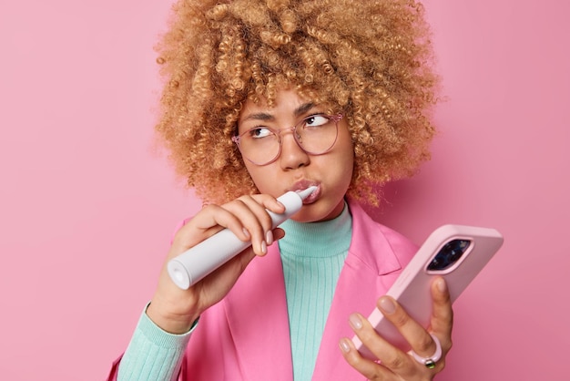 Ernstige doordachte krullende vrouw poetst tanden met elektrische tandenborstel en tandpasta ondergaat tandverzorgingsroutine gebruikt mobiele telefoon om te chatten draagt een bril geïsoleerd over roze achtergrond