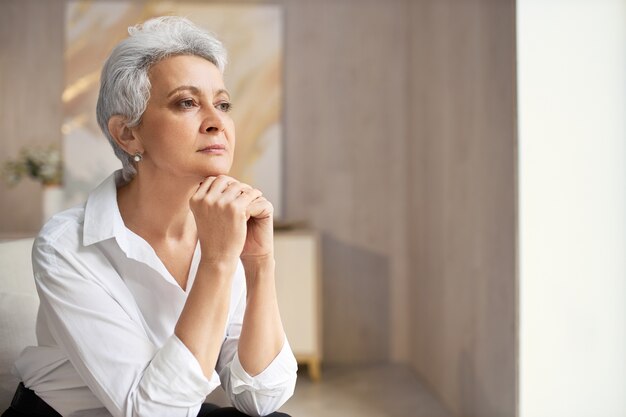 Ernstige elegante gepensioneerde vrouw met kort kapsel poseren binnenshuis met handen onder de kin, wegkijken met peinzende gezichtsuitdrukking, nadenken over een idee of beslissing