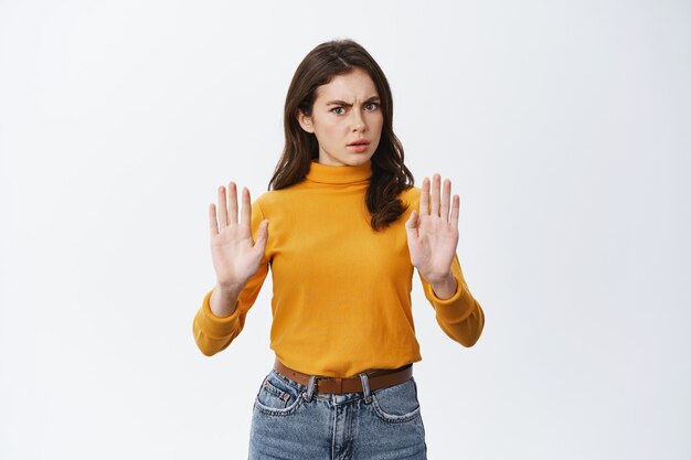 Ernstige en vastberaden vrouw zegt nee, toont verbied en verbiedt gebaar, vraagt om te kalmeren of iets slechts te weigeren, fronsend, staande op een witte muur