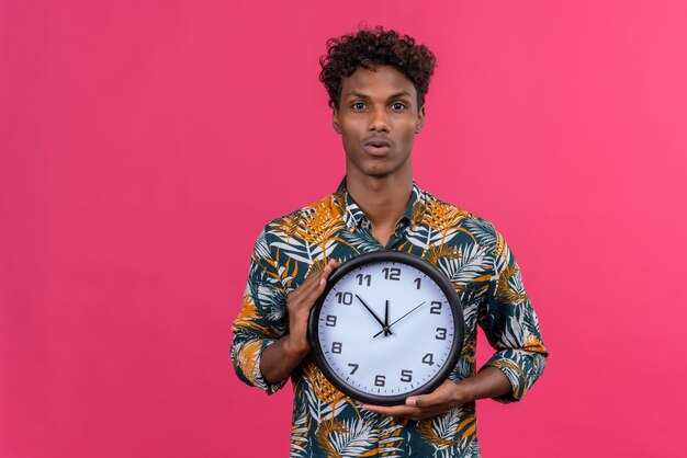 Ernstige en verwarde jonge knappe donkerhuidige man met krullend haar in bladeren bedrukt overhemd met wandklok op een roze achtergrond