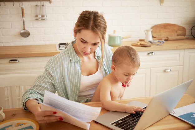 Ernstige geconcentreerde jonge vrouw papieren in handen studeren, rekeningen online betalen, zittend aan de keukentafel voor open laptop met zoontje op haar schoot. Klein kind te typen op draagbare computer