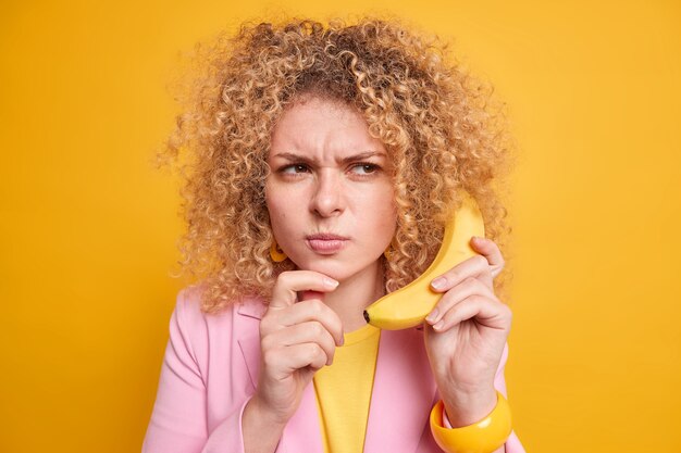 Ernstige gekrulde Europese vrouw met ontevredenheidsuitdrukking doet alsof ze een belangrijk gesprek heeft, houdt een smakelijke banaan in de buurt van het oor, formeel gekleed grijnzend gezicht geïsoleerd over gele muur.