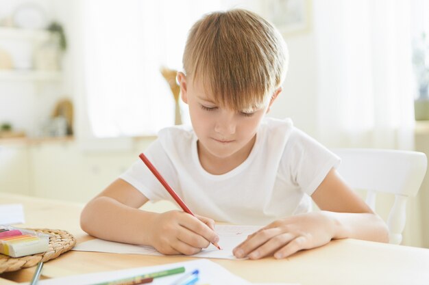 Ernstige gerichte schooljongen in wit t-shirt vermaakt zichzelf binnenshuis met behulp van rood potlood tekenen of schetsen aan houten tafel geïsoleerd tegen stijlvolle woonkamer