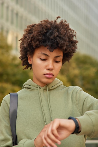 Ernstige gezonde vrouw met krullend haar kijkt naar smartwatch controleert tijd of fitnessresultaten draagt hoodie wandelingen buiten in stedelijke omgeving tegen onscherpe achtergrond. atletische vrouw houdt de voortgang in de gaten