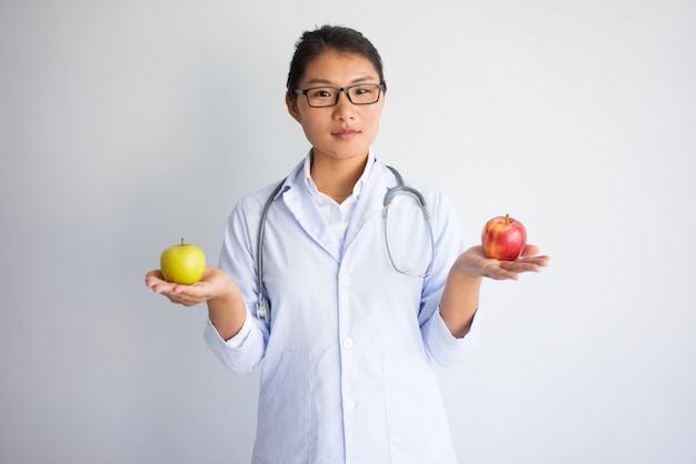 Ernstige jonge Aziatische vrouwelijke arts die rode en gele appel houdt.