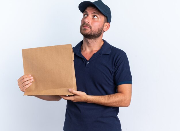 Ernstige jonge blanke bezorger in blauw uniform en pet met papieren pakket op zoek naar geïsoleerd op een witte muur