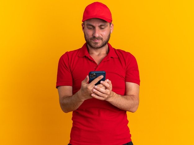 Ernstige jonge blanke bezorger in rood uniform en pet die mobiele telefoon vasthoudt en bekijkt geïsoleerd op oranje muur met kopieerruimte