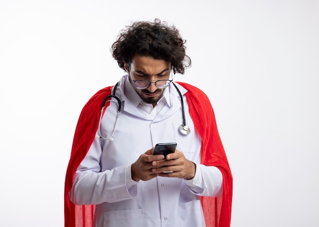 Ernstige jonge blanke superheld man in optische bril dragen dokter uniform met rode mantel en met stethoscoop om nek houdt en kijkt naar telefoon