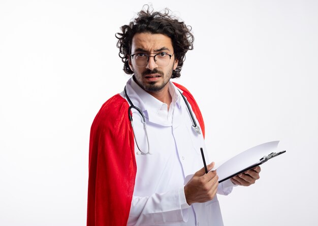 Ernstige jonge blanke superheld man in optische bril dragen dokter uniform met rode mantel en met stethoscoop om nek houdt potlood en klembord