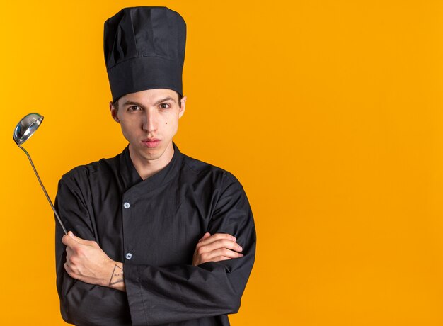 Ernstige jonge blonde mannelijke kok in chef-kok uniform en pet staande met gesloten houding met pollepel kijken camera geïsoleerd op oranje muur met kopieerruimte