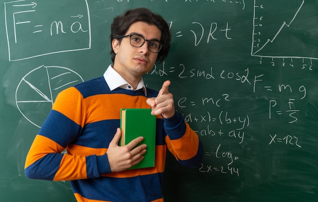 Ernstige jonge kaukasische meetkundeleraar met een bril die voor het bord staat in de klas met een boek