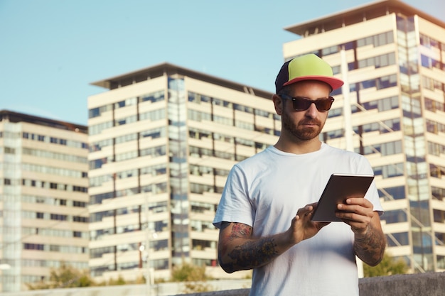 Ernstige jonge man met een wit effen t-shirt en een rode, gele en zwarte vrachtwagenchauffeurhoed die naar zijn tablet kijkt tegen stadsgebouwen en lucht