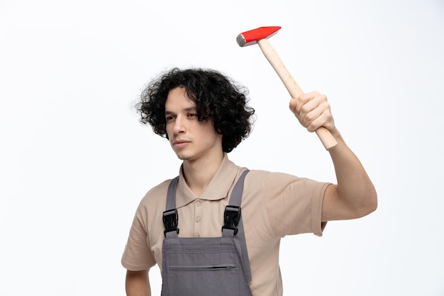 Ernstige jonge mannelijke bouwvakker die een uniform draagt en naar de opstaande hamer kijkt die omhoog is geïsoleerd op een witte achtergrond