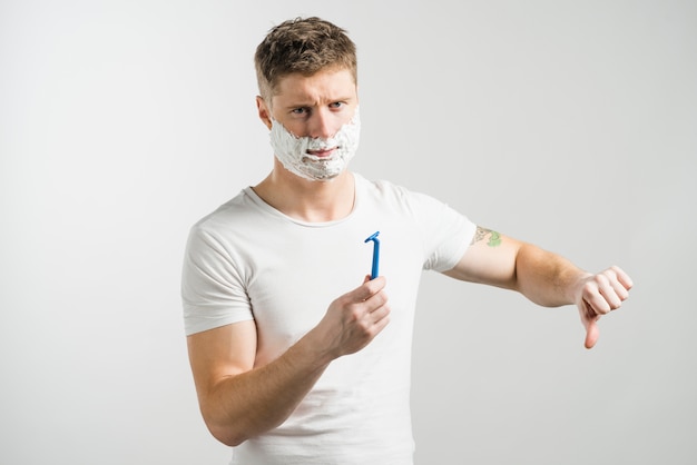 Ernstige jonge mens met het scheren van schuim op zijn gezicht die blauw scheermes houden die in hand duimen tonen neer tegen grijze achtergrond