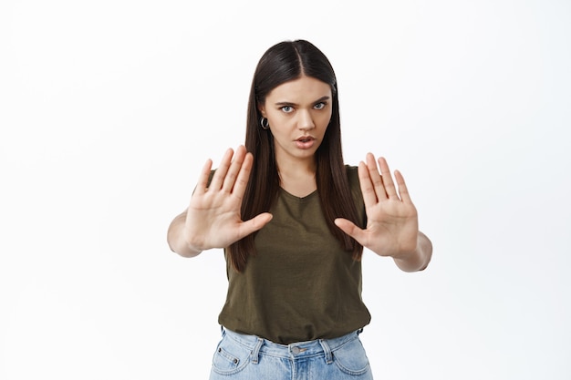 Ernstige jonge vrouw strekt haar handen uit om te blokkeren, iemand te stoppen, nee te zeggen, tegen een witte muur te staan, iets slechts te weerspiegelen, witte muur