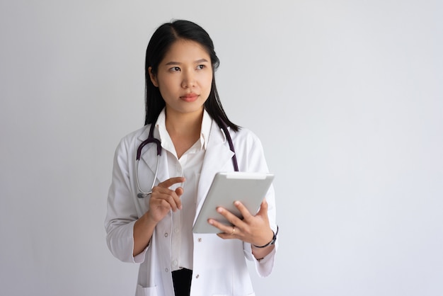 Ernstige jonge vrouwelijke arts die tabletcomputer met behulp van