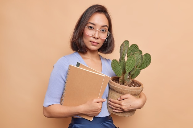 Ernstige jonge vrouwelijke leraar kijkt zelfverzekerd naar voren draagt optische bril blauwe trui houdt twee notitieblokken en ingemaakte cactus geïsoleerd over beige muur