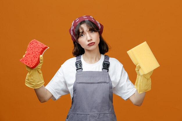Ernstige jonge vrouwelijke schoonmaker met uniforme rubberen handschoenen en bandana die naar de camera kijkt met sponzen geïsoleerd op een oranje achtergrond