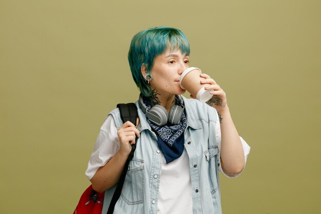 Ernstige jonge vrouwelijke student met koptelefoon en bandana op nek en rugzak die rugzak grijpt en naar de zijkant kijkt terwijl hij koffie drinkt uit een papieren koffiekopje geïsoleerd op olijfgroene achtergrond