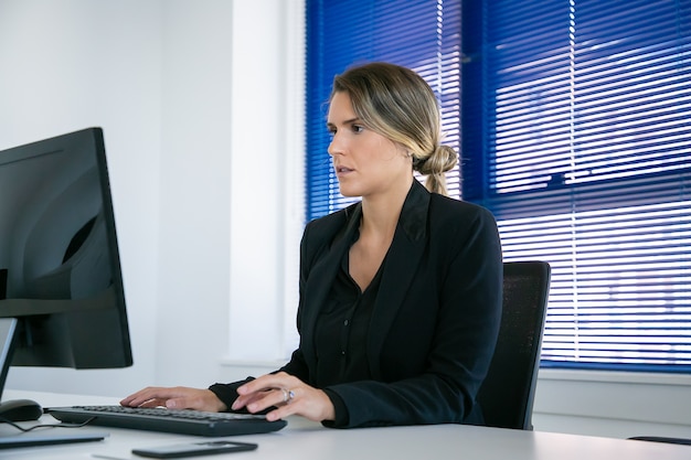 Ernstige jonge zakenvrouw jas dragen, computer gebruiken op de werkplek op kantoor, typen en kijken naar display. gemiddeld schot. digitaal communicatieconcept