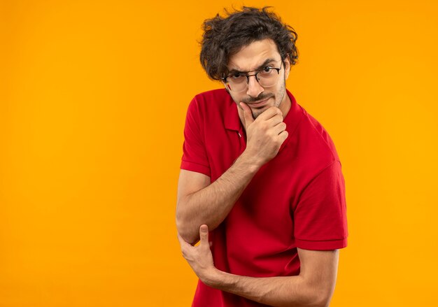 Ernstige jongeman in rood shirt met optische bril legt hand op kin en kijkt geïsoleerd op oranje muur