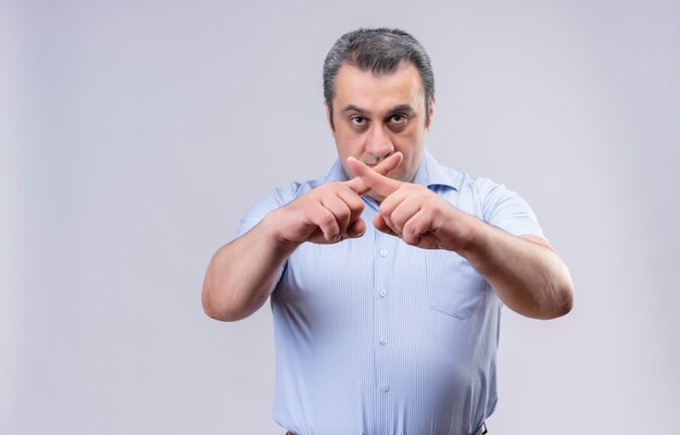 Ernstige man van middelbare leeftijd die een blauw overhemd draagt dat de vorm van het x-teken met zijn vingers op een witte achtergrond toont