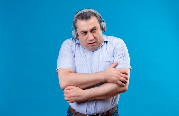 Ernstige man van middelbare leeftijd met een blauw verticaal gestreept shirt in een koptelefoon die het koud heeft terwijl hij probeert warm te blijven op een blauwe achtergrond
