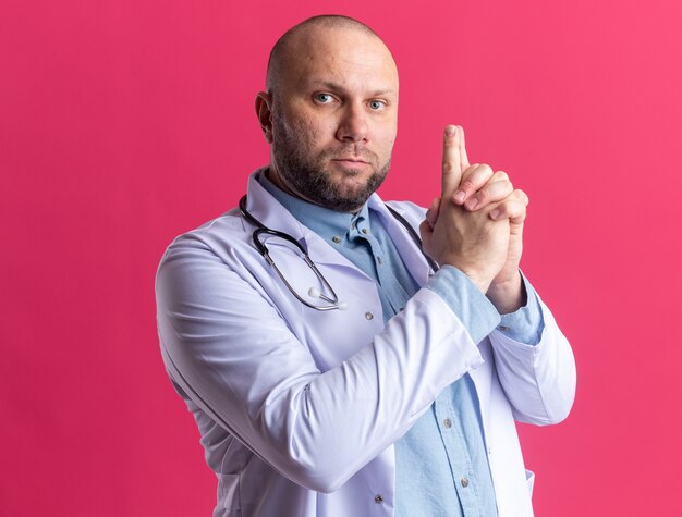 Ernstige mannelijke arts van middelbare leeftijd met een medisch gewaad en een stethoscoop die naar de voorkant kijkt en een pistoolgebaar doet dat op een roze muur is geïsoleerd