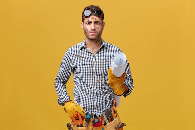 Ernstige mannelijke bouwer met riem van instrumenten die geruit overhemd, beschermende brillen en handschoenen dragen die documenten in hand houden die over gele muur worden geïsoleerd. mensen, reparatie en constructie