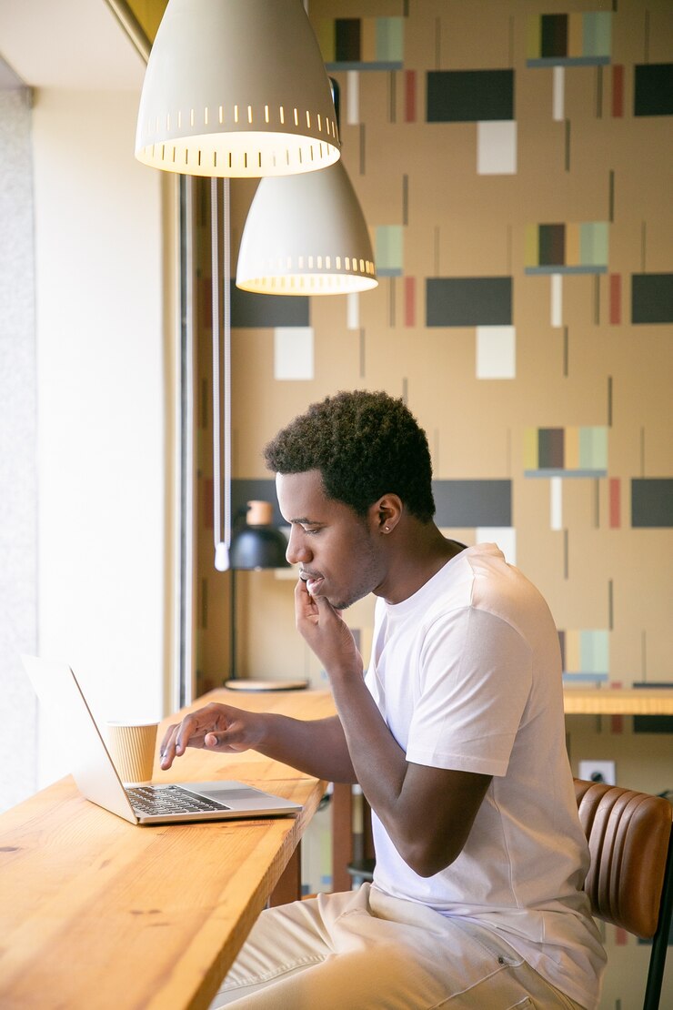 Gratis Foto ernstige mannelijke freelance die op laptop werkt en op cel in co-workingruimte spreekt