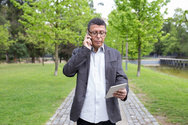 Ernstige mens gebruikend tablet en sprekend op telefoon in park