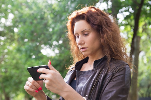 Ernstige mooie dame die op smartphone in stadspark doorbladeren